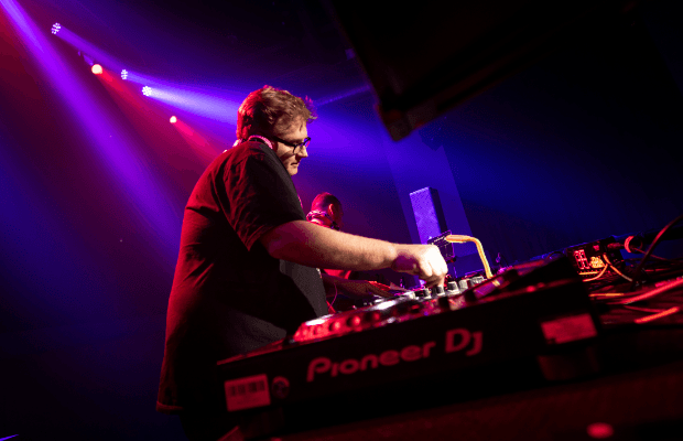 D-Zero wird von der Seite gezeigt und agiert am Mischpult. Der Hintergrund ist bunt und zeigt eine typische Disco-Atmosphäre. Der DJ wird von hinten mit einem Strahler angeleuchtet.