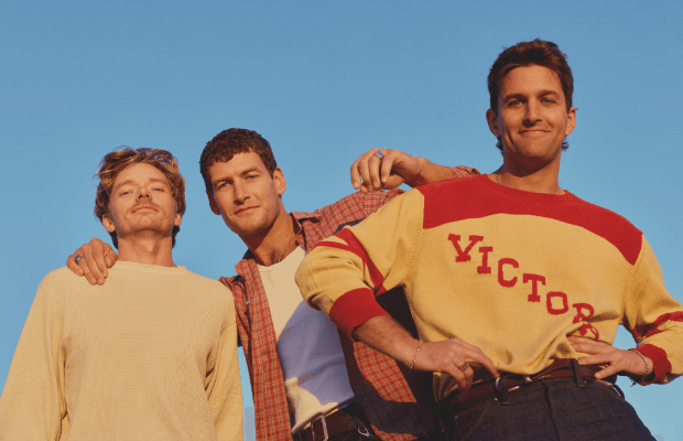 Aufnahme der drei Musiker der Band Sons of the East, die sich in den Armen liegen, von schräg unten gegen einen hellblauen Himmel.