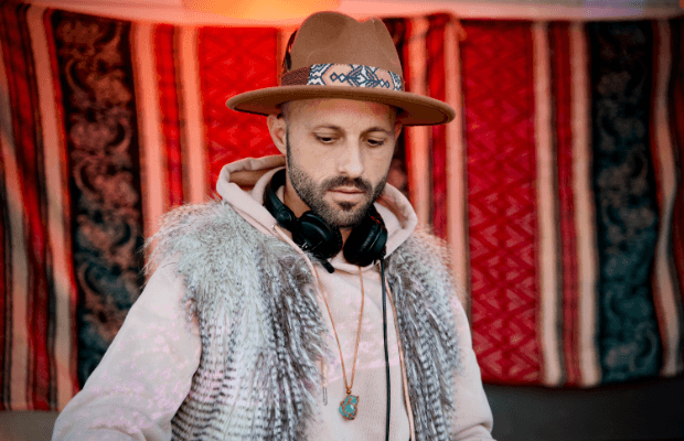 DJ Tor trägt einen braunen Hut und Kopfhörer um den Hals. Er steht vor einem rot-blauen Stoffhintergund und blickt konzentriert nach unten.