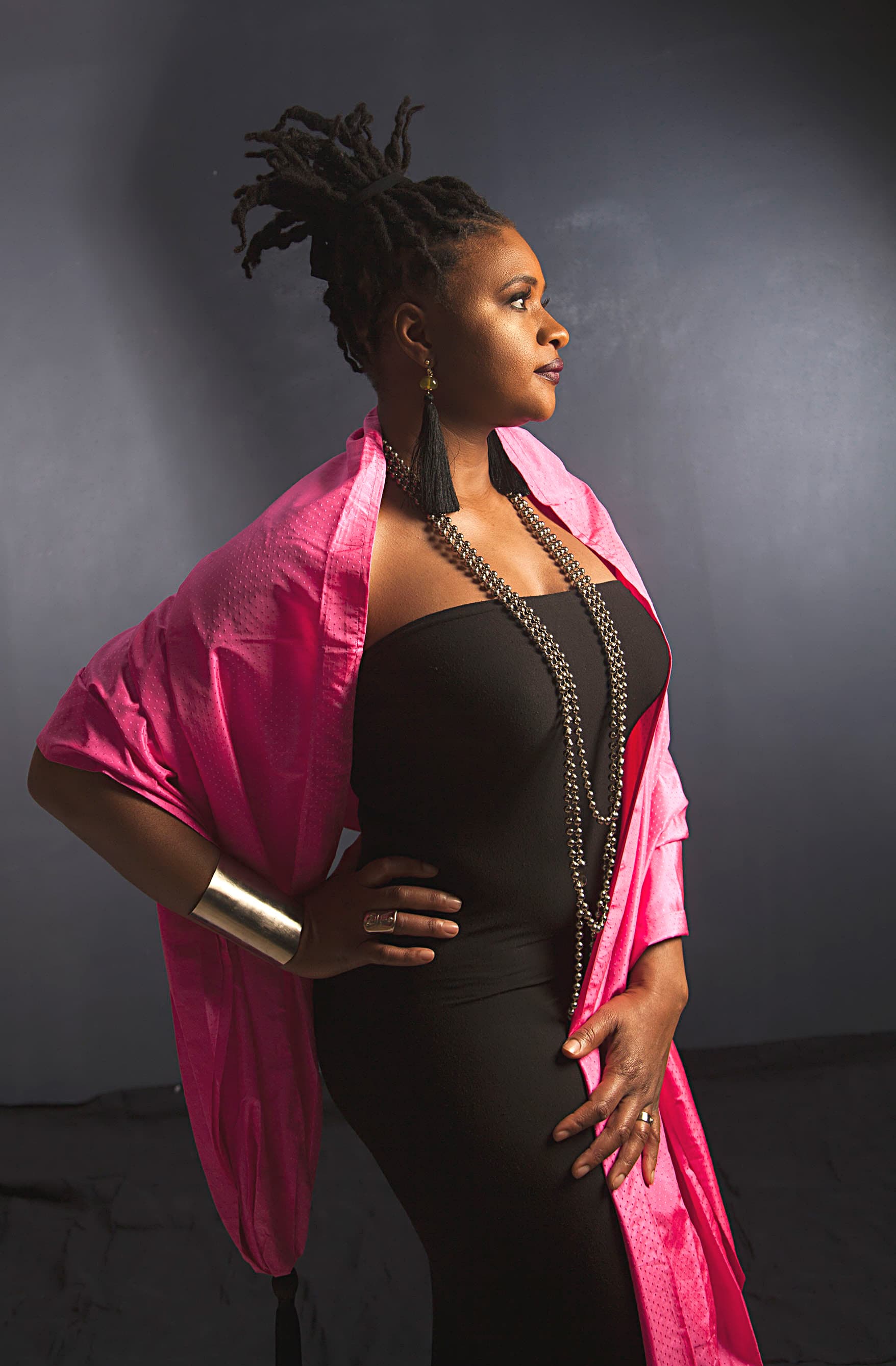 Die Künstlerin Lizz Wright steht seitlich zur Kamera. Sie trägt ihr Haar in einem hohen Zopf, ein schwarzes schulterfreies Oberteil, ein leuchtend pinkes Tuch über den Schultern und eine auffällige silberne Kette.