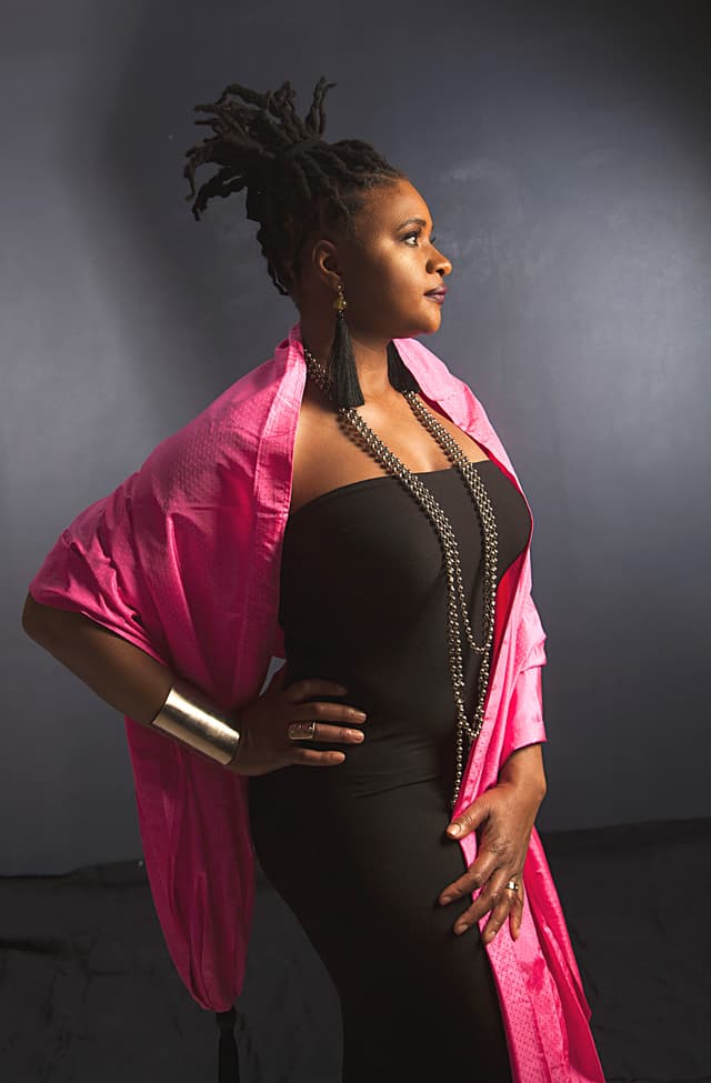 Die Künstlerin Lizz Wright steht seitlich zur Kamera. Sie trägt ihr Haar in einem hohen Zopf, ein schwarzes schulterfreies Oberteil, ein leuchtend pinkes Tuch über den Schultern und eine auffällige silberne Kette.