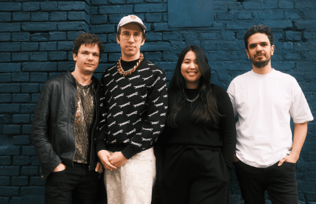 Die drei Musiker und eine Musikerin stehen vor einer dunkelgrün gestrichenen Steinwand und blicken in die Kamera.