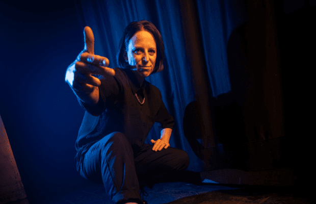 Die DJane Emma Mc sitzt in der Hocke und macht eine Handbewegung in Richtung Kamera. Der Hintergrund ist blau-schwarz gehalten.