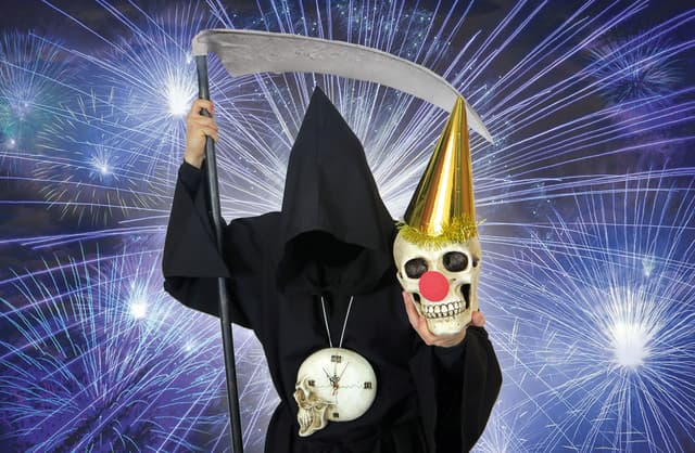 Als Sensenmann verkleideter Mensch, das Gesicht ist nicht erkennbar, mit Clownsnase und Partyhut, hält eine Totenkopfmütze, vor einem Feuerwerks-Hintergrund.