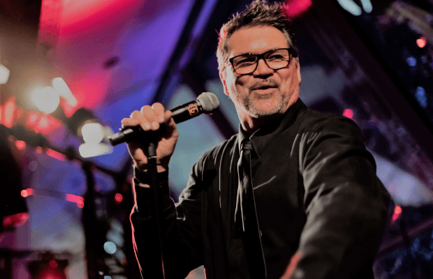 Der Sänger Joe Whitney mit Brille und schwarzem Outfit singt mit Mikrofon auf einer beleuchteten Bühne.