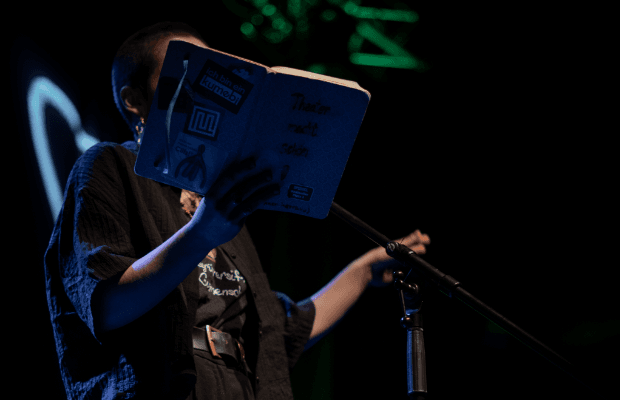 Eine Frau steht in dunkler Bühnenatmosphäre vor einem Mikrofon und hält ein aufgeschlagenese Buch in der Hand. Das Buch verdeckt ihr Gesicht.