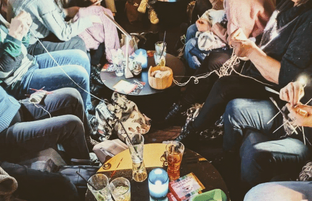 Man sieht die Unterkörper einiger Menschen, die sich in zwei Reihen gegenüber sitzen und gemeinsam stricken. Auf zwei Tischen in der Mitte stehen Getränke.