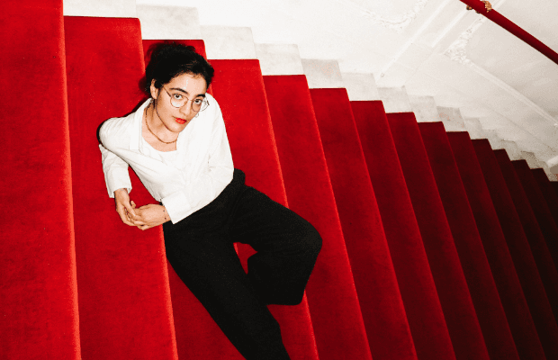 In Draufsicht aufgenommenes Foto der Comedienne Ana Lucia, wie sie auf einer mit rotem Samt bezogenen Treppe liegt, rotes Geländer an einer Wand mit Stuckverziehungen. Sie trägt eine Brille, weiße Bluse, schwarze Hosen und Stiefel.