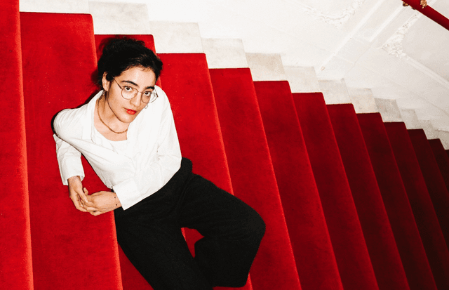 In Draufsicht aufgenommenes Foto der Comedienne Ana Lucia, wie sie auf einer mit rotem Samt bezogenen Treppe liegt, rotes Geländer an einer Wand mit Stuckverziehungen. Sie trägt eine Brille, weiße Bluse, schwarze Hosen und Stiefel.