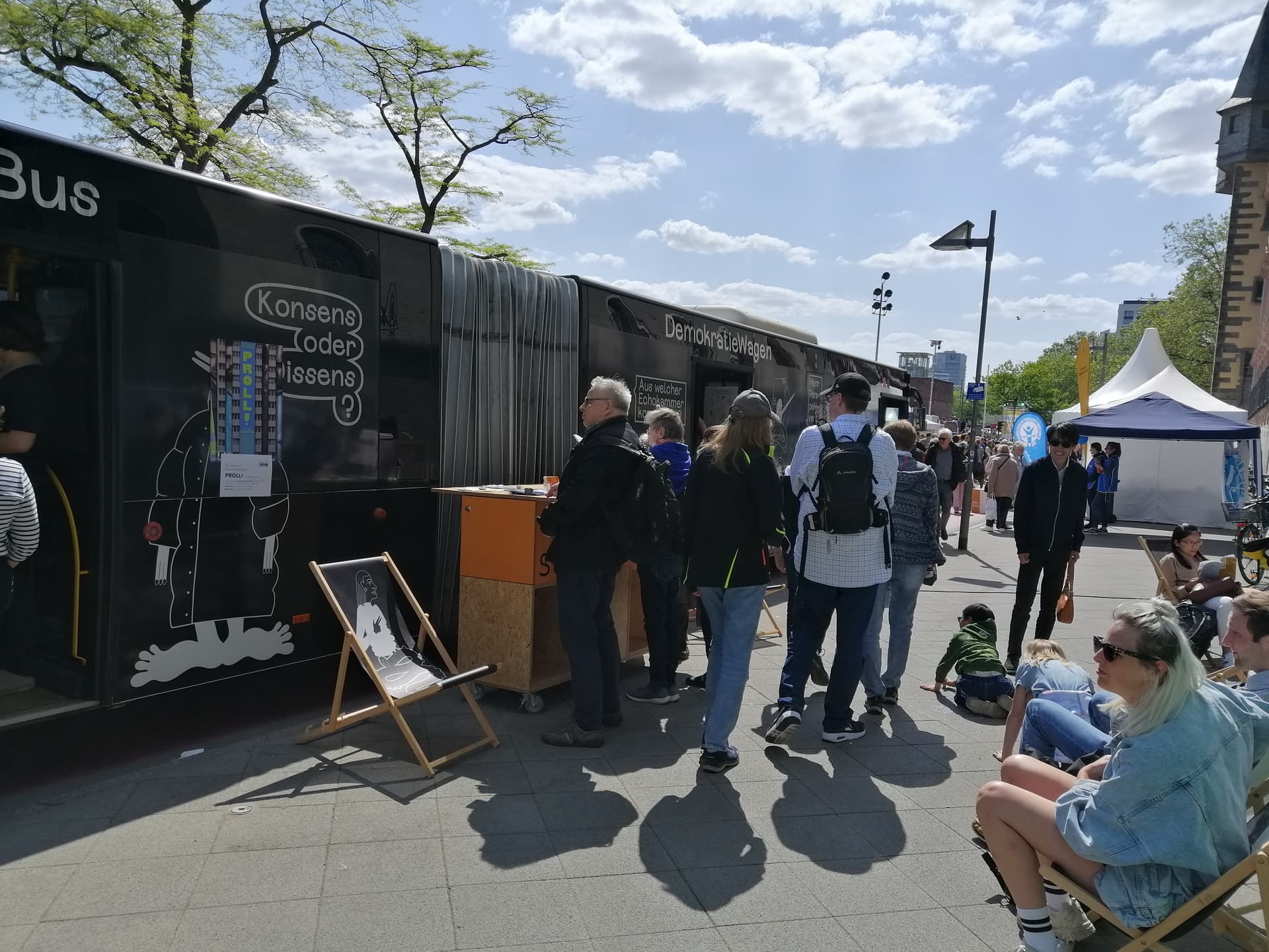 Mehrere Menschen stehen bei sonnigem Wetter in einer Schlange vor einem schwarzen Bus mit Schriftzügen und Illustrationen auf einem öffentlichen Platz; einige Personen sitzen daneben.