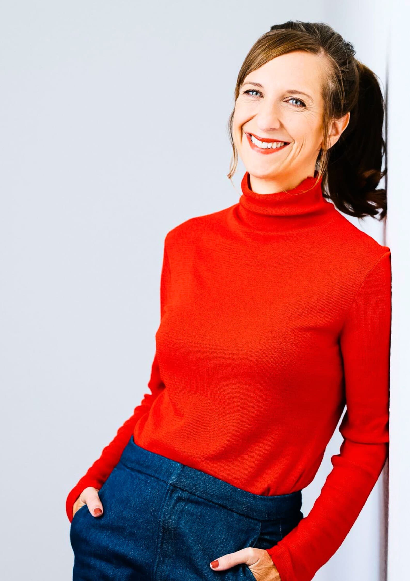 Porträt der Musikerin mit rotem Rollkragenpullover und rot geschminkten Lippen, die herzlich lachend in die Kamera blickt.
