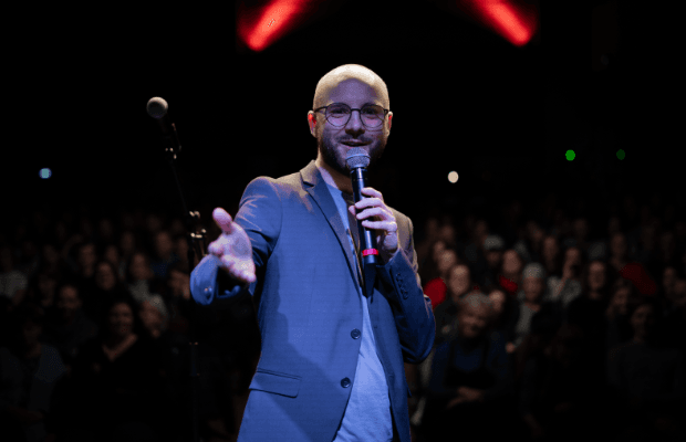 Der Moderator Finn Holitzka steht auf einer Bühne, streckt dem Fotografen lächelnd die rechte Hand entgegen, in der linken hält er ein Mikrofon. Verschwommen sieht man das Publikum im Hintergrund.