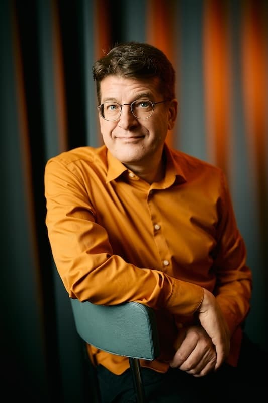 Porträt des Comedians Nils Heinrich in orangenem Hemd und mit Brille, der freundlich in die Kamera blickt.