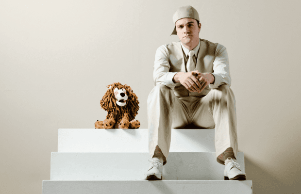 Künstler im beigen Anzug mit Krawatte und Schirmmütze, auf einer weißen, dreistufigen Treppe sitzend, neben einem Kuscheltier-Löwen.