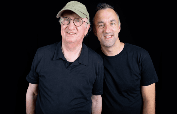 Porträtaufnahme zweier Männer in schwarzen T-Shirts vor schwazem Hintergrund. Sie lächeln in die Kamera, der linke Mann trägt eine khakigrüne Schirmmütze und eine Brille mit runden Gläsern.