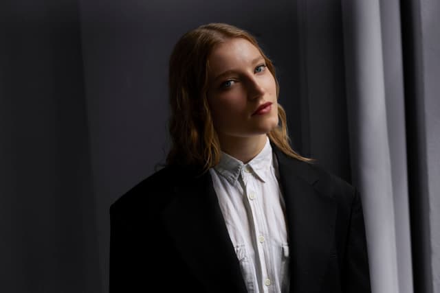 Porträt der Jazzmusikerin Emma Rawicz: sie steht vor einem dunklen Hintergrund und blickt leicht nach rechts, trägt ein schwarzes Sakko über einem weißen Hemd, ihr blondes Haar fällt locker über die Schultern.