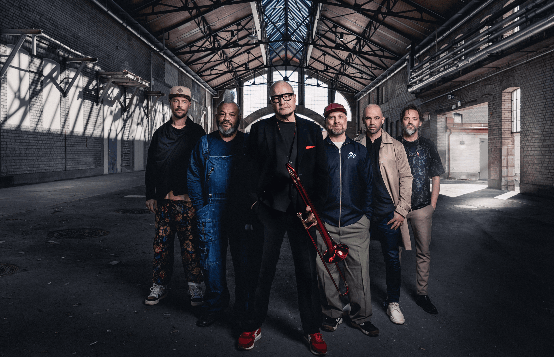 Die sechs Musiker der Nils Landgrend Funk Unit stehen nebeneinander in einer großen Industriehalle mit Backsteinwänden und Glasdach. In der Mitte Nils Landgrend in dunklem Anzug mit rotem Einstecktuch und hält eine rote Posaune, die anderen Bandmitglieder tragen legere Kleidung und blicken in die Kamera.
