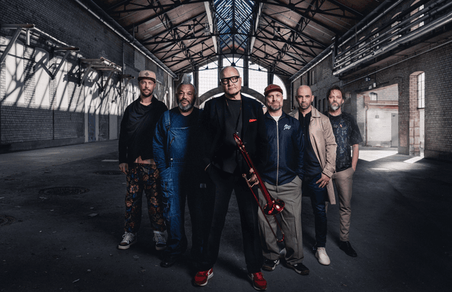 Die sechs Musiker der Nils Landgrend Funk Unit stehen nebeneinander in einer großen Industriehalle mit Backsteinwänden und Glasdach. In der Mitte Nils Landgrend in dunklem Anzug mit rotem Einstecktuch und hält eine rote Posaune, die anderen Bandmitglieder tragen legere Kleidung und blicken in die Kamera.