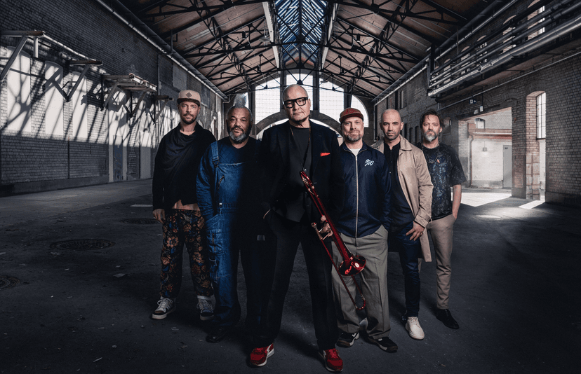 Die sechs Musiker der Nils Landgrend Funk Unit stehen nebeneinander in einer großen Industriehalle mit Backsteinwänden und Glasdach. In der Mitte Nils Landgrend in dunklem Anzug mit rotem Einstecktuch und hält eine rote Posaune, die anderen Bandmitglieder tragen legere Kleidung und blicken in die Kamera.