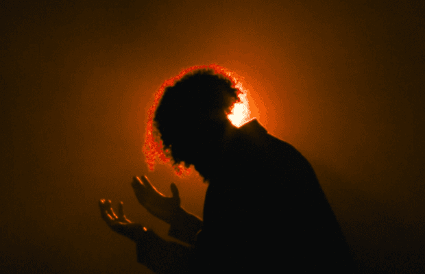 Silhouette einer Person mit erhobenen Händen vor einem warmen, orangefarbenem Licht. Das Gegenlicht erzeugt einen leuchtenden Rand um die lockigen Haare.