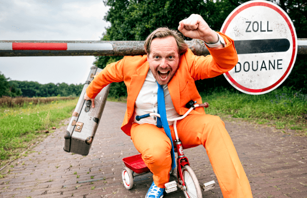 Patrick Nederkoorn trägt einen orangefarbenen Anzug mit blauer Krawatte und sitzt auf einem Dreirad. Er fährt damit unter einer Zoll-Schranke hindurch, reckt die linke Hand zur Faust und hält einen Koffer in der rechten. Der Mund steht im offen, als juble oder schreie er.
