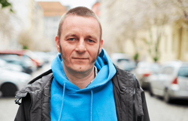 Proträt des Autors, mit verschwommener Straßenszene im Hintergrund, der direkt in die Kamera blickt und einen hellblauen Kapuzenpulli unter einer schwarzen Jacke trägt.