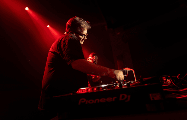 D-Zero wird von der Seite gezeigt und agiert am Mischpult. Der Hintergrund ist bunt und zeigt eine typische Disco-Atmosphäre. Der DJ wird von hinten mit einem Strahler angeleuchtet.