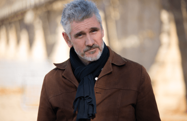 Porträt des Musikers Curtis Stigers mit brauner Lederjacke und schwarzem Schal vor sonnig, verschwommenem Hintergrund.