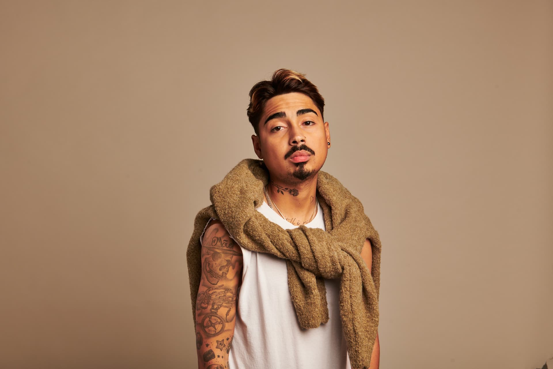 Porträt des Comedy-Artists Tony Bauer vor beigem Hintergrund, mit ärmellosem weißen Shirt, Pullover über den Schultern und sichtbaren Tattoos.