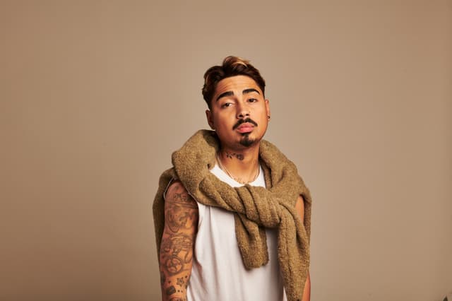 Porträt des Comedy-Artists Tony Bauer vor beigem Hintergrund, mit ärmellosem weißen Shirt, Pullover über den Schultern und sichtbaren Tattoos.