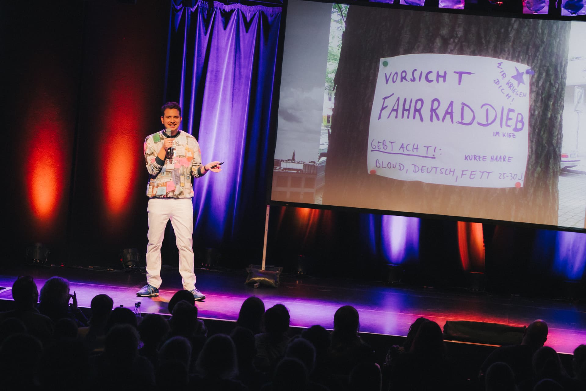 Joab Nist steht auf einer beleuchtete Bühne und hält ein Mikrofon in derHand, während hinter ihm ein Foto mit einem handgeschriebenen Zettel ("Vorsicht Fahrraddieb. gebt Acht! Kurze Haare. Blond, deutsch, fett") projiziert wird; im Vordergrund ist das Publikum zu sehen.