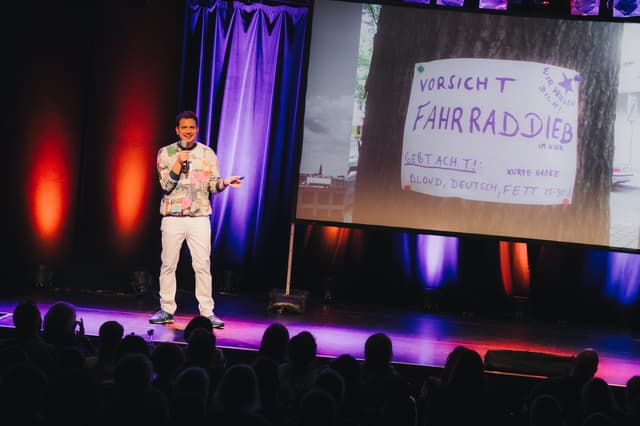 Joab Nist steht auf einer beleuchtete Bühne und hält ein Mikrofon in derHand, während hinter ihm ein Foto mit einem handgeschriebenen Zettel ("Vorsicht Fahrraddieb. gebt Acht! Kurze Haare. Blond, deutsch, fett") projiziert wird; im Vordergrund ist das Publikum zu sehen.