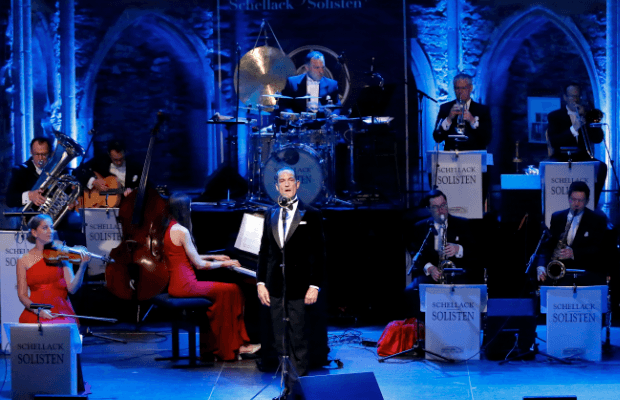 Der Sänger Denis Wittberg im schwarzen Frack steht am Mikrofon vor dem Jazz-Ensemble Schellack-Solisten auf einer blau beleuchteten Bühne.