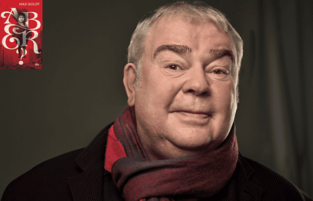 Porträt des Autors Max Goldt mit kurzen grauen Haaren. Er trägt einen dunklen Mantel und einen rot gemusterten Schal. Neutrale, dunkle Hintergrundfläche, ruhiger Gesichtsausdruck, Blick leicht zur Kamera.