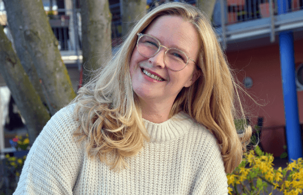 Das Foto zeigt eine blonde Frau. Sie trägt eine Brille und neigt den Kopf leicht zur rechten Bildseite. Im Hintergrund erahnt man einen Baum, einen gelb blühenden Busch und ein buntes Gebäude.