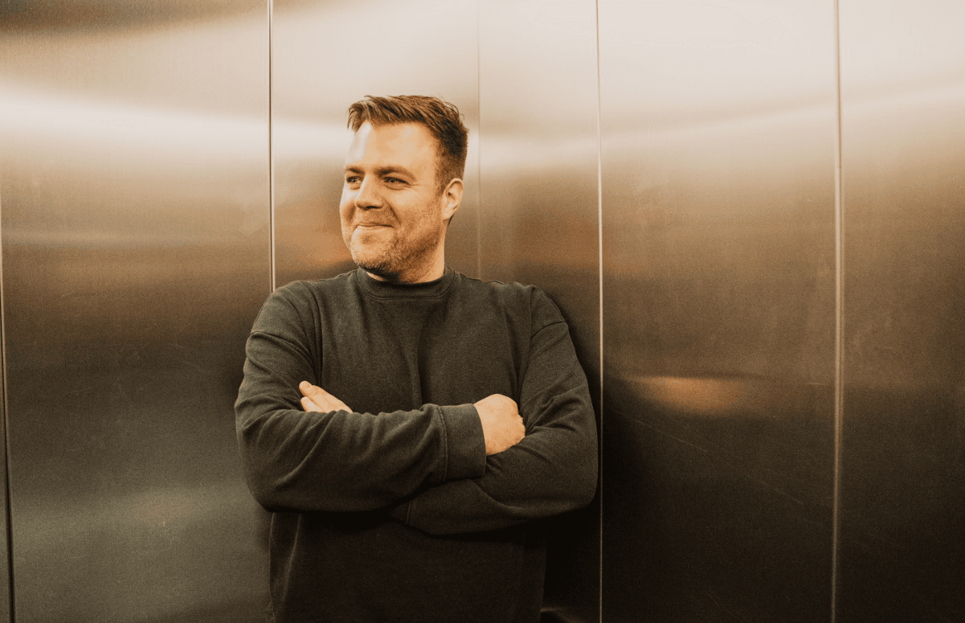 Ein Mann mit kurzen, hellbraunen Haaren und dunklem Pullover steht mit verschränkten Armen vor einer metallisch glänzenden Wand und blickt lächelnd zur Seite.