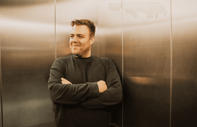 Ein Mann mit kurzen, hellbraunen Haaren und dunklem Pullover steht mit verschränkten Armen vor einer metallisch glänzenden Wand und blickt lächelnd zur Seite.