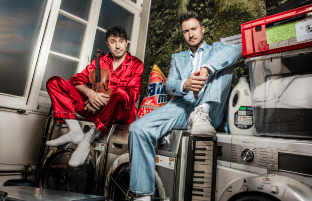 Zwei Männer in schrillen Anzühen sitzen auf Waschmaschinen, einer hat eine Gege im Arm, der andere sein Bein auf ein Keyboard gestellt.