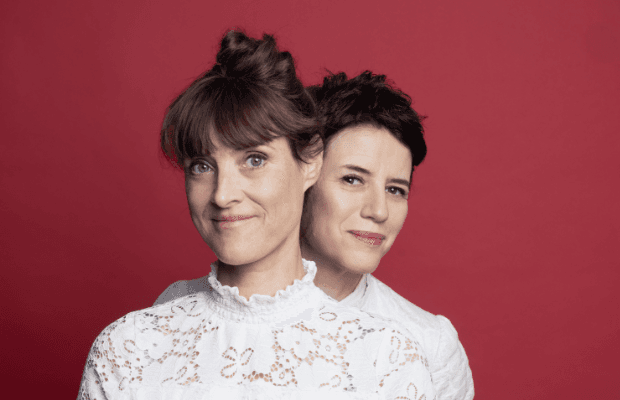 Die beiden Musikerinnen stehen hintereinander vor einem Weinroten Hintergrund, die eine blickt der anderen lächelnd über die Schulter, beidet tragen weiße Blusen und blicken lächelnd in die Kamera.