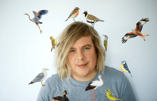 Ein Porträt Dominik Eulbergs vor hellem Hintergrund mit illustrierten Vögeln untertschiedlichster Arten, die scheibar um ihn herumfliegen, auf seinem Kopf und seinen Schultern sitzen.